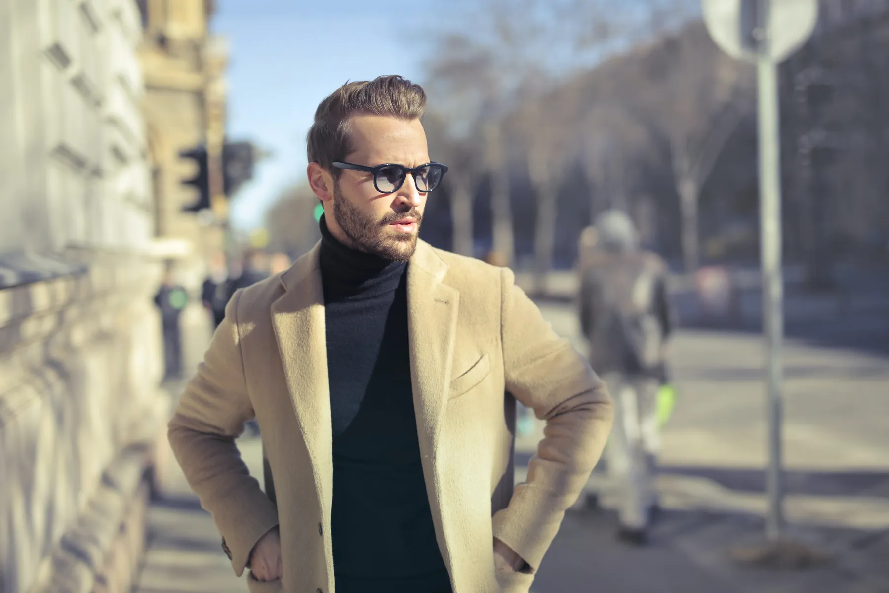 A confident man in a camel coat and glasses on a city street